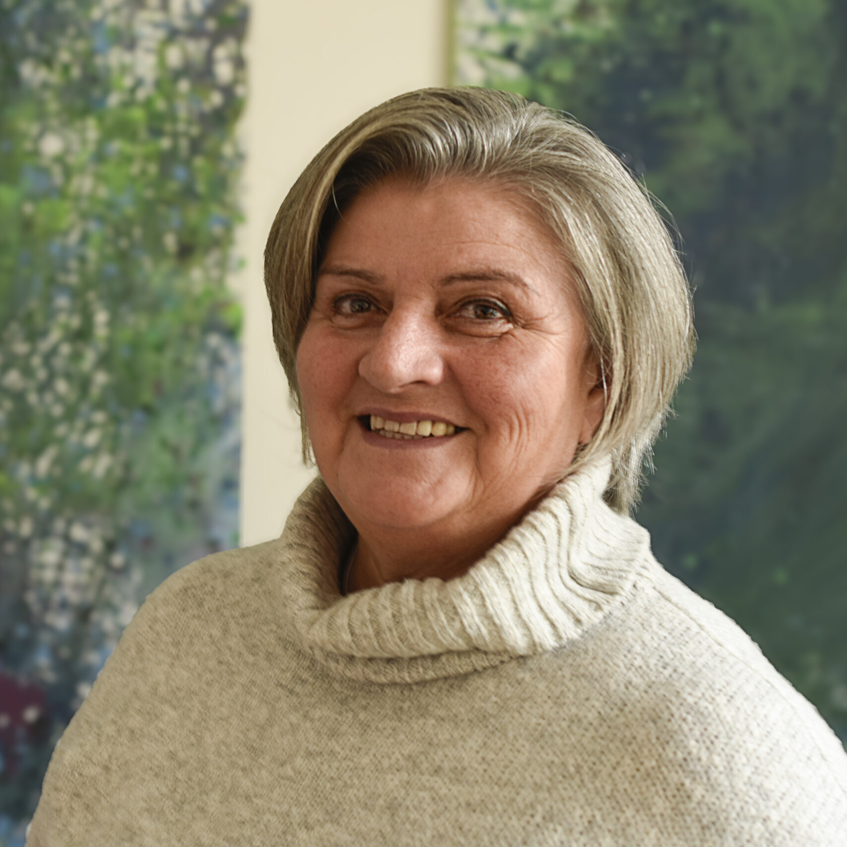 Eine Frau mit kurzen grauen Haaren und einem cremefarbenen Rollkragenpullover lächelt vor einem abstrakten Kunstwerk in Grün und Blau. Eine Frau mit kurzen grauen Haaren und einem cremefarbenen Rollkragenpullover lächelt vor einem abstrakten Kunstwerk in Grün und Blau.