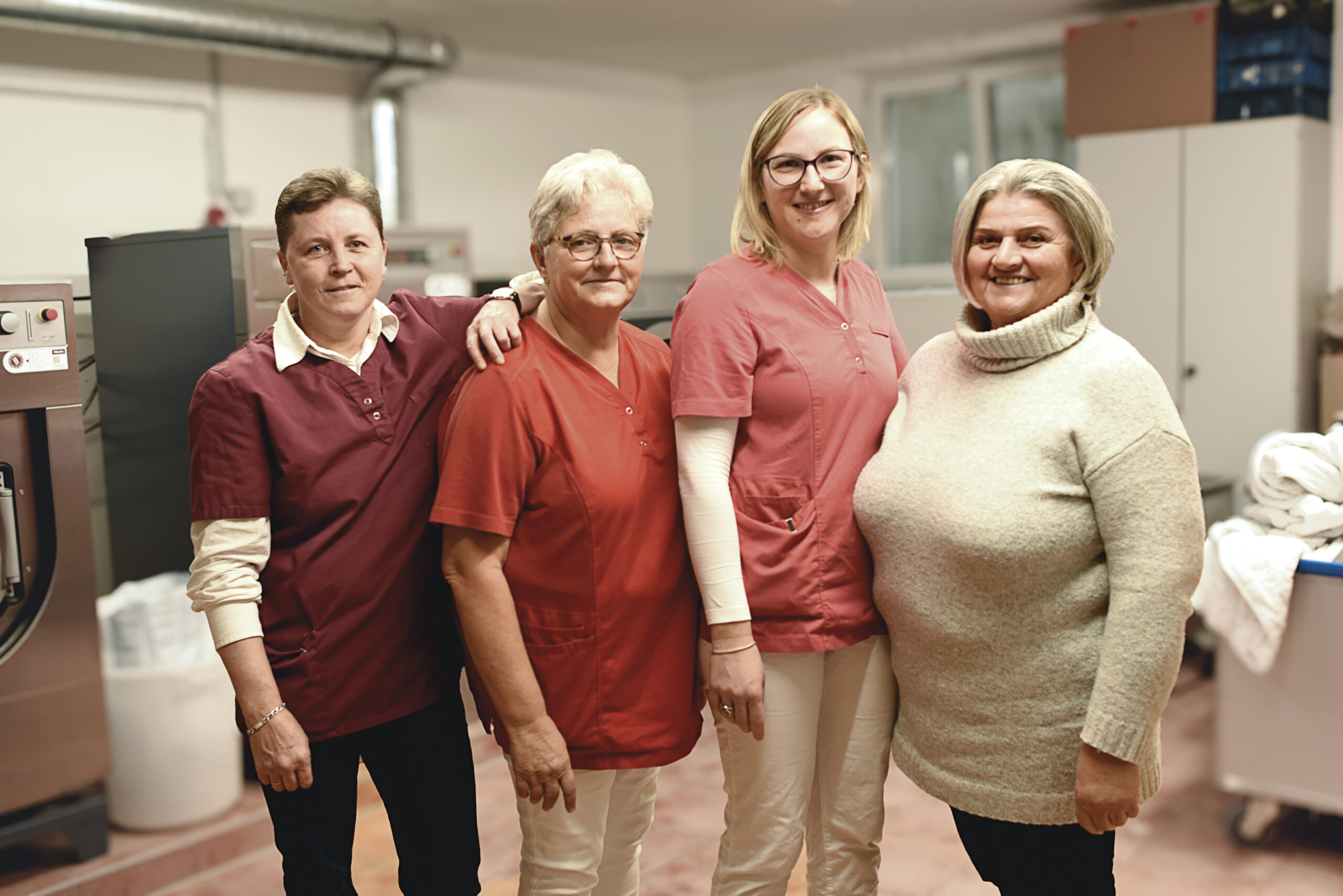 Vier Frauen stehen zusammen in einer Wäscherei oder einem Arbeitsraum, drei tragen rote Uniformen und eine einen beigen Pullover; alle lächeln in die Kamera. Vier Frauen stehen zusammen in einer Wäscherei oder einem Arbeitsraum, drei tragen rote Uniformen und eine einen beigen Pullover; alle lächeln in die Kamera.