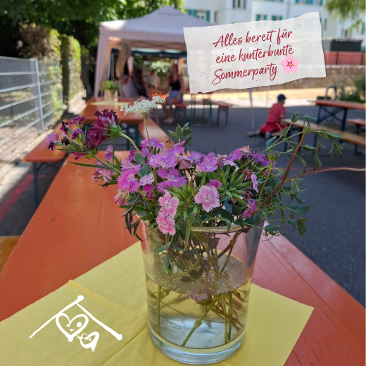 Auf einem orangefarbenen Tisch im Freien steht ein Glasgefäß mit violetten Blumen auf einer gelben Serviette. Im Hintergrund versammeln sich Menschen unter einem Zelt. Der deutsche Text lautet: „Alles bereit für eine bunte Sommerparty.“.