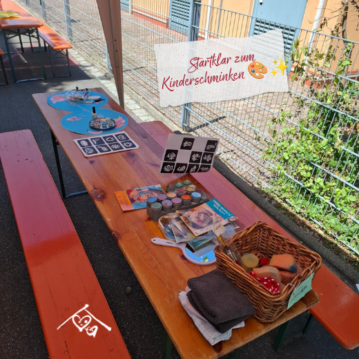 Ein Tisch im Freien mit Gesichtsfarben, Pinseln, einem Spiegel, einem Korb mit Schwämmen und einem Blatt mit Motiven für das Kinderschminken. Die Bildunterschrift lautet „Startklar zum Kinderschminken“ und ist mit Emojis für Kunst und Glitzer versehen.