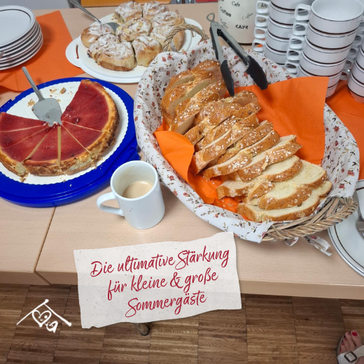 Ein Tisch mit Kuchen, geschnittenem Brot, einer Tasse Kaffee, gestapelten Tellern und Tassen. Auf einem Schild in deutscher Sprache steht: „Die ultimative Stärkung für kleine & große Sommergäste“.