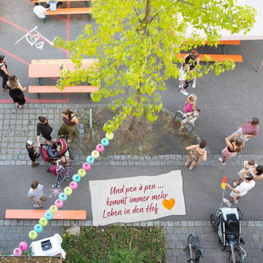 Im Innenhof versammeln sich Menschen unter einem Baum um Tische und Dekorationen. In der Nähe stehen Fahrräder und Kinderwagen. Der deutsche Text lautet: „Und peu à peu… kommt immer mehr Leben in den Hof“, ergänzt durch ein orangefarbenes Herz.