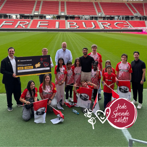 Eine Gruppe von Kindern und Erwachsenen steht auf einem Fußballplatz im Freiburger Stadion und hält Fahnen sowie einen großen Spendenscheck über 2.000 Euro in den Händen. Der deutsche Text lautet: Jede Spende zählt!, ergänzt durch Herz- und Fußball-Strichzeichnungen. Weibliche Inspiration. Stark für Frauen. Stark für Menschen.