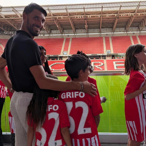 Ein Mann steht mit Kindern in roten Fußballtrikots mit der Aufschrift GRIFO 32 da und blickt auf ein leeres Stadion mit roten Sitzplätzen und einem grünen Fußballfeld.
