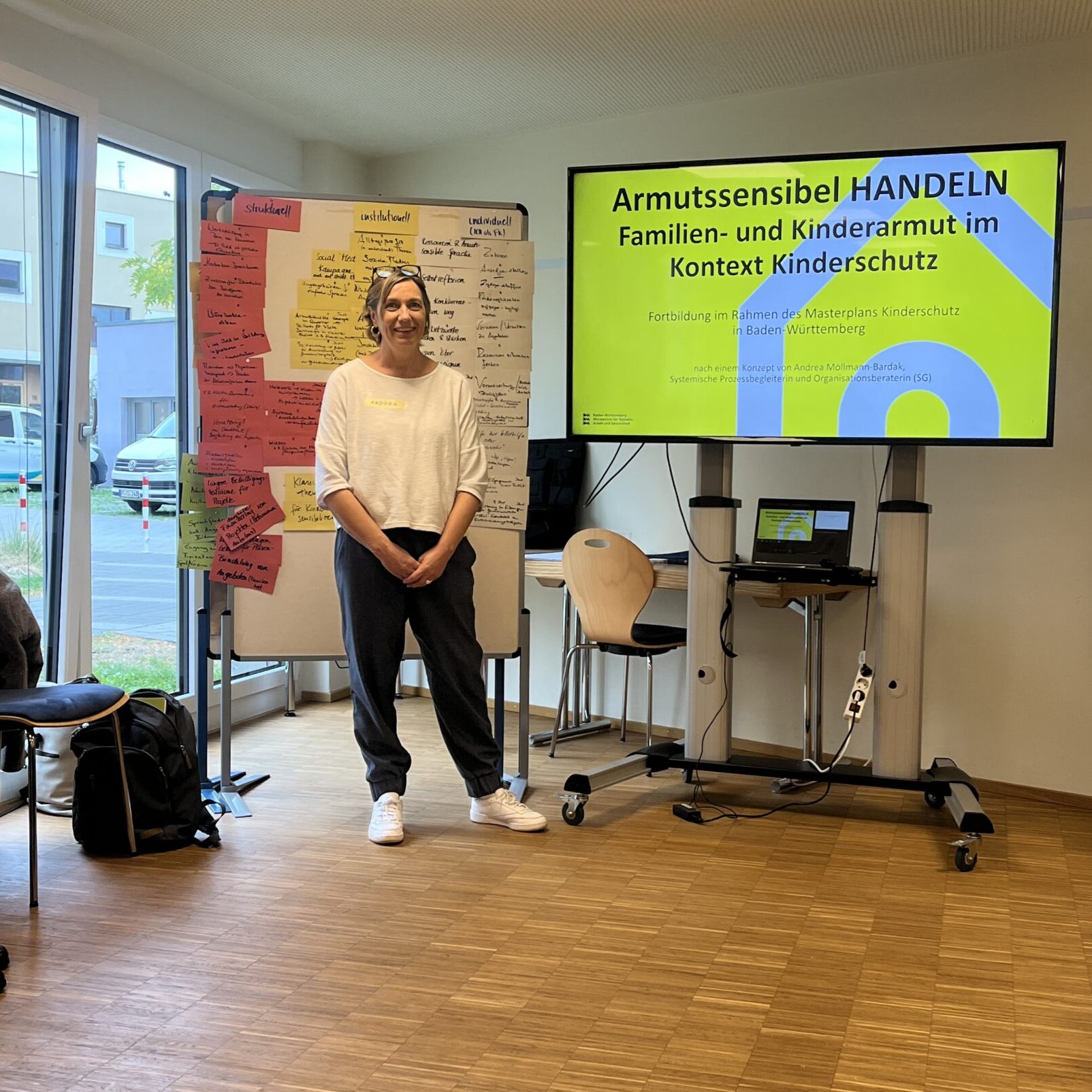 Eine Frau steht vor einer Präsentationswand mit der Aufschrift Armutssensibel HANDELN neben einem Whiteboard, das mit Haftnotizen übersät ist.