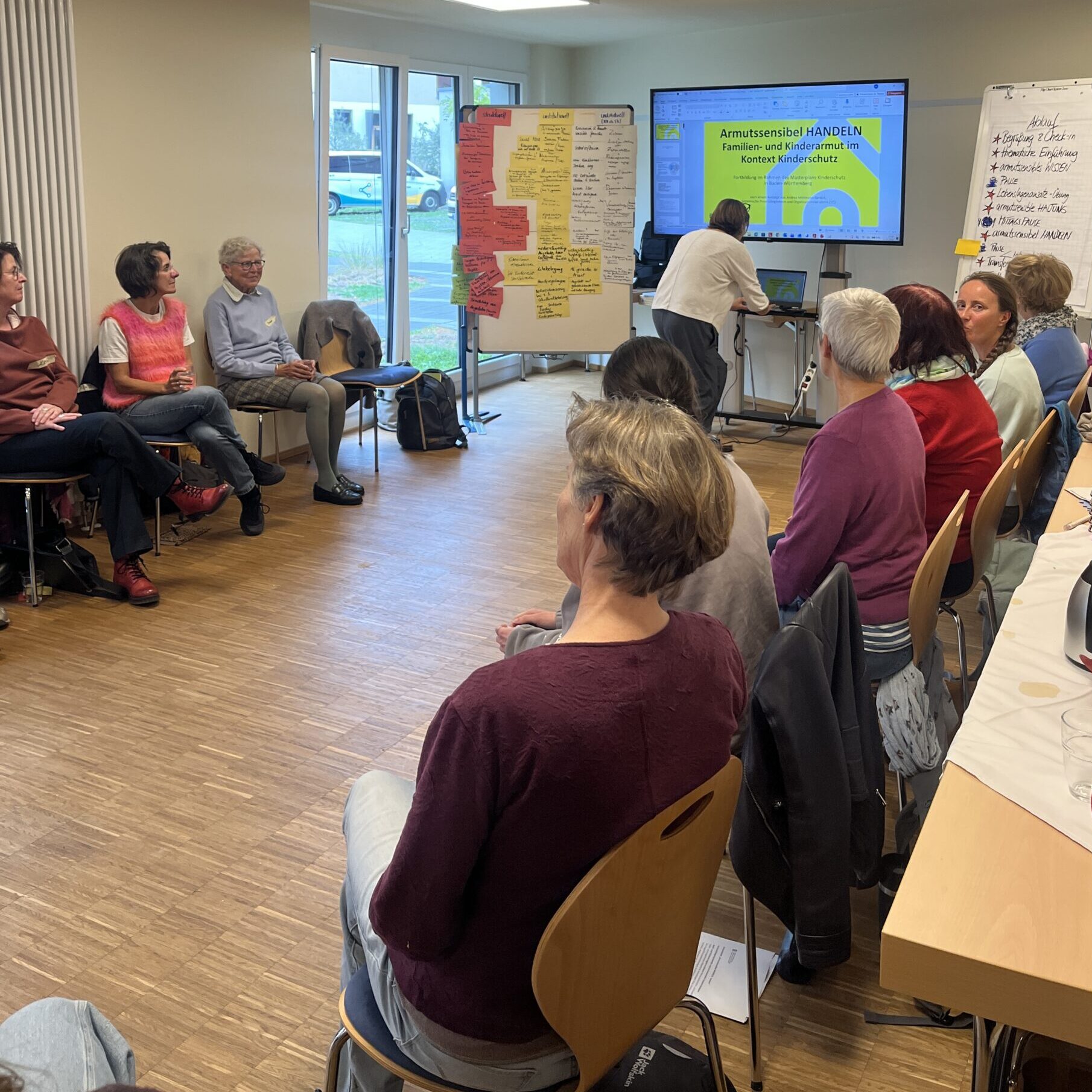 Eine Gruppe von Menschen sitzt in einem Besprechungsraum im Kreis und blickt auf eine Leinwand und ein Whiteboard, die für eine Präsentation zum Thema Kinderschutz genutzt werden.