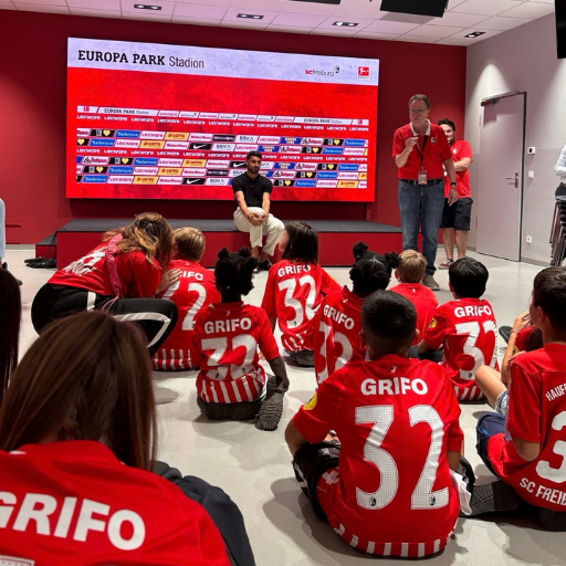 Eine Gruppe von Kindern in roten Grifo 32-Trikots sitzt auf dem Boden und blickt zu einem Mann, der vor einer großen roten Leinwand im Europa-Park-Stadion sitzt, während Erwachsene daneben stehen.