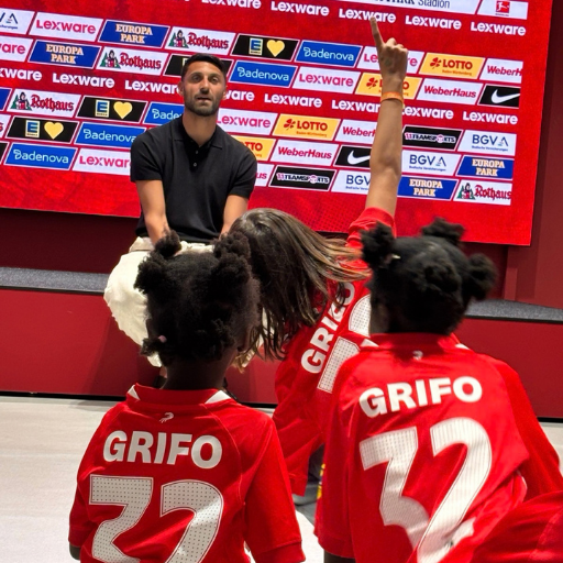 Ein Mann sitzt vor einer Gruppe von Kindern, die rote GRIFO-32-Trikots tragen. Ein Kind hebt die Hand, während im Hintergrund ein roter Vorhang mit zahlreichen Sponsorenlogos zu sehen ist.