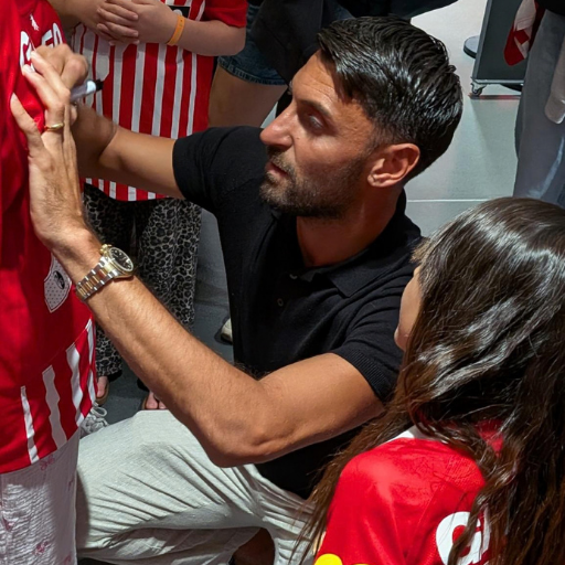 Ein Mann kniet nieder und signiert ein rot-weiß gestreiftes Sporttrikot, das ein Kind trägt, während ein anderes Kind in einem ähnlichen Trikot daneben steht und zusieht.