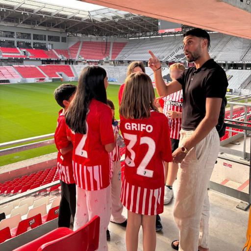 Ein Mann spricht zu einer Gruppe von Kindern in rot gestreiften Fußballtrikots, die in einem Stadion mit leeren Sitzplätzen stehen, während im Hintergrund ein grüner Rasen zu sehen ist.